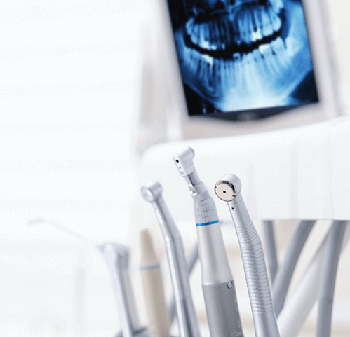 Close-up of various dental handpieces, including drills and other instruments, in a dental clinic setting. In the blurred background, a screen displays a dental X-ray image of a patient's teeth and jaw
