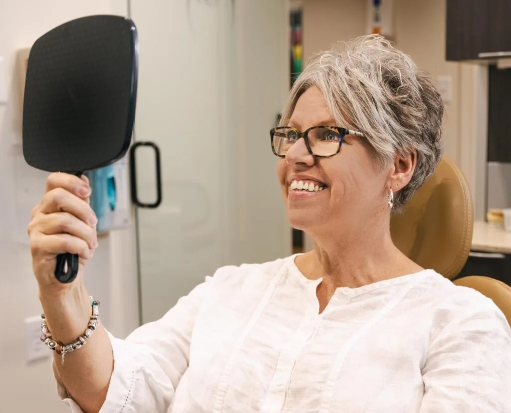 An older woman with short gray hair and glasses is sitting in a dental chair, holding a black handheld mirror and smiling as she examines her teeth. She is wearing a white blouse and a beaded bracelet. The background shows a dental office setting with cabinets and equipment.