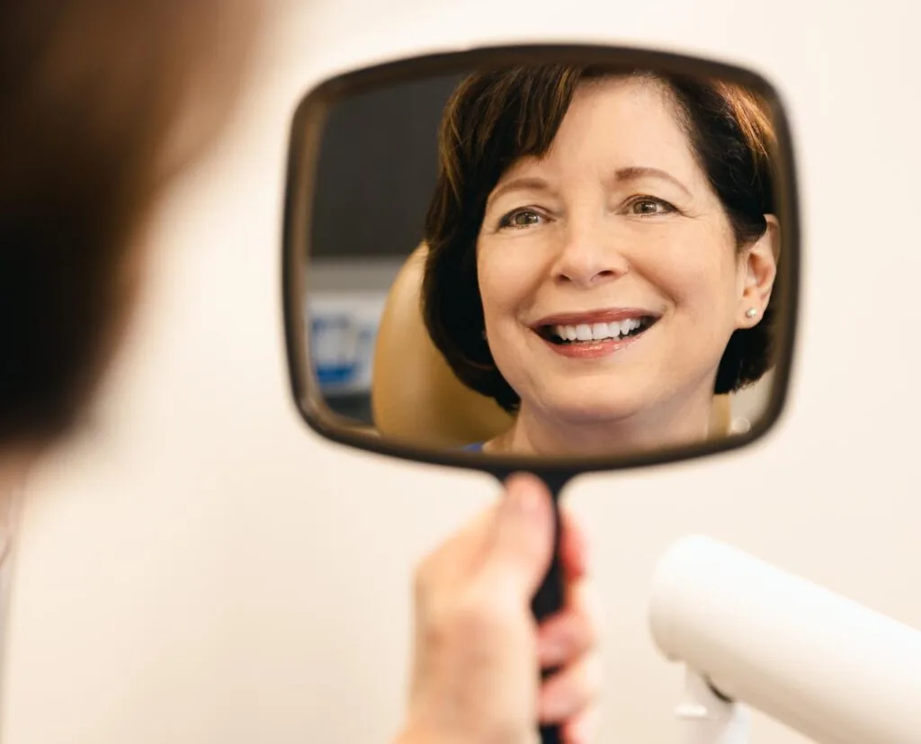A woman with short dark hair is smiling while looking at her reflection in a handheld mirror at a dental clinic. She appears happy and satisfied with her teeth, possibly after a dental procedure. The background is blurred, showing a dental chair and clinic setting.
