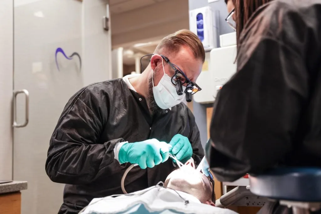 A male dentist wearing magnifying loupes, a face mask, and green gloves performs a dental procedure on a patient lying in a dental chair. An assistant in black scrubs is partially visible, assisting with the treatment.
