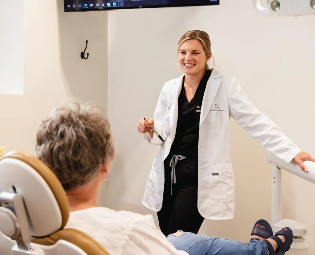 A female dentist wearing a white coat over black scrubs is smiling and engaging in conversation with a patient seated in a dental chair. She is holding a dental tool in one hand while leaning against a support. The patient, an older woman with short gray hair, is attentively listening. The background features a dental office setting with a screen mounted on the wall.