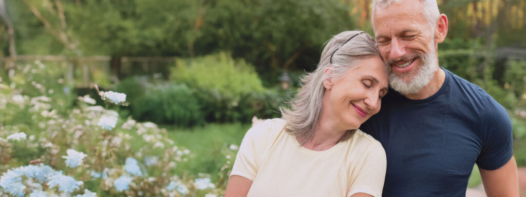 An older couple embraces in a lush garden, smiling. The woman, wearing a light yellow shirt, leans her head on the man’s shoulder with her eyes closed, while the man, in a navy blue shirt, smiles with his eyes slightly closed. Soft sunlight filters through the greenery and blooming flowers in the background.